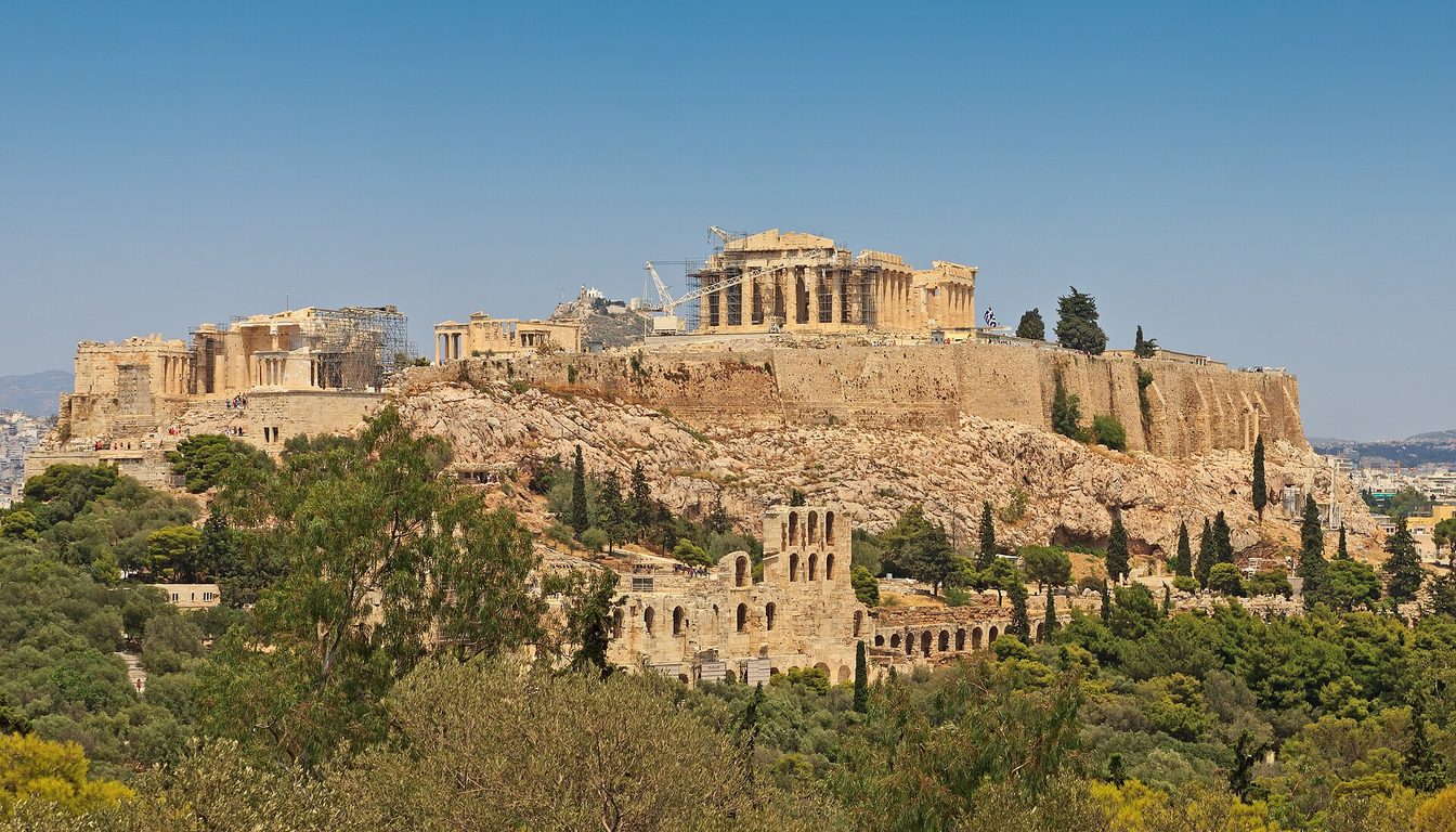 Acropolis hill with the Parthenon and Odeon of Herodes Atticus rising above green trees under a clear blue sky