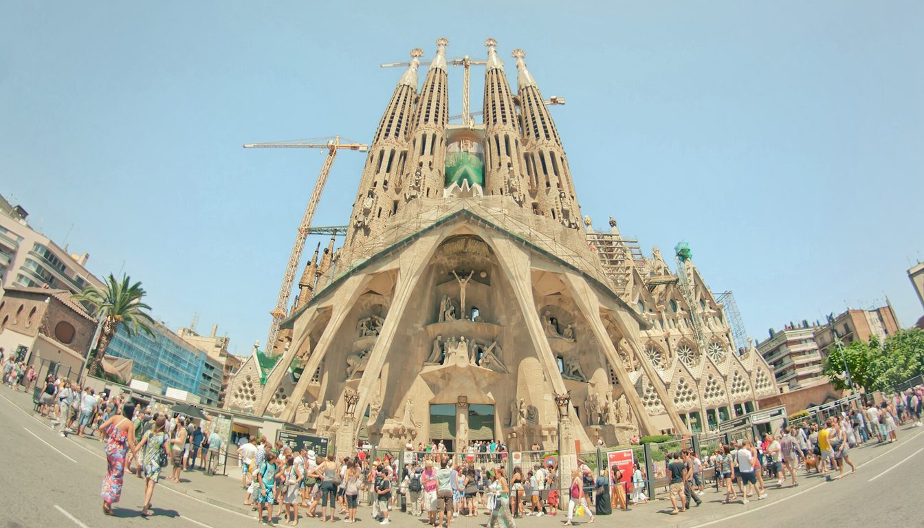 Crowds of tourists gathered in front of the Passion façade of the Sagrada Família under a clear blue sky