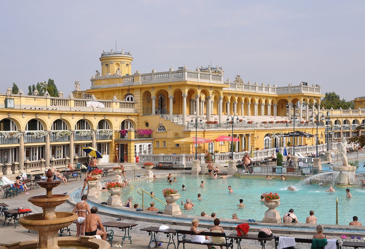 Bathers in the outdoor pool in front of the yellow neo-baroque Széchenyi Baths building under a blue sky