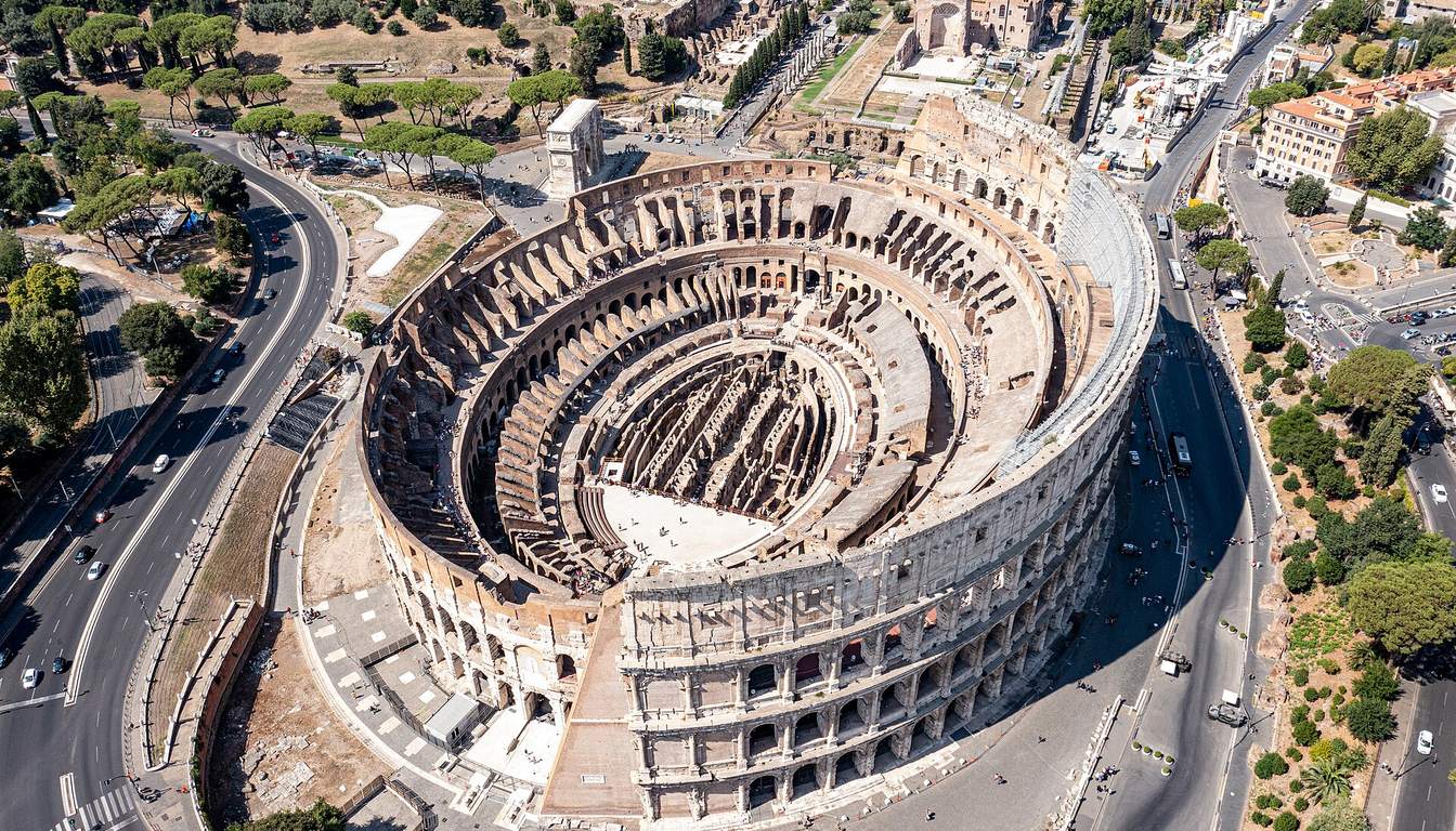 Drone view of the elliptical Colosseum with its exposed interior, surrounded by roads, pine trees and Rome's buildings