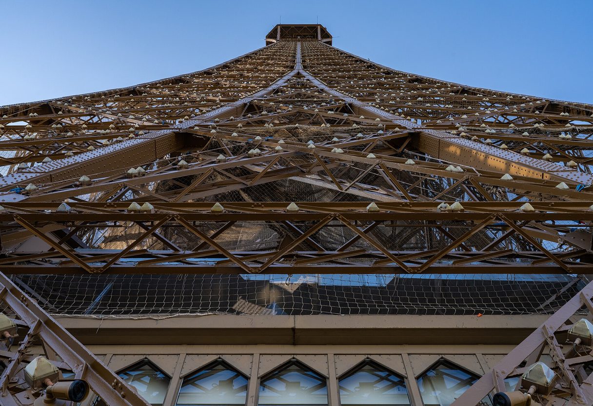 View up the Eiffel Tower ironwork from the second-floor platform toward the summit under a blue sky