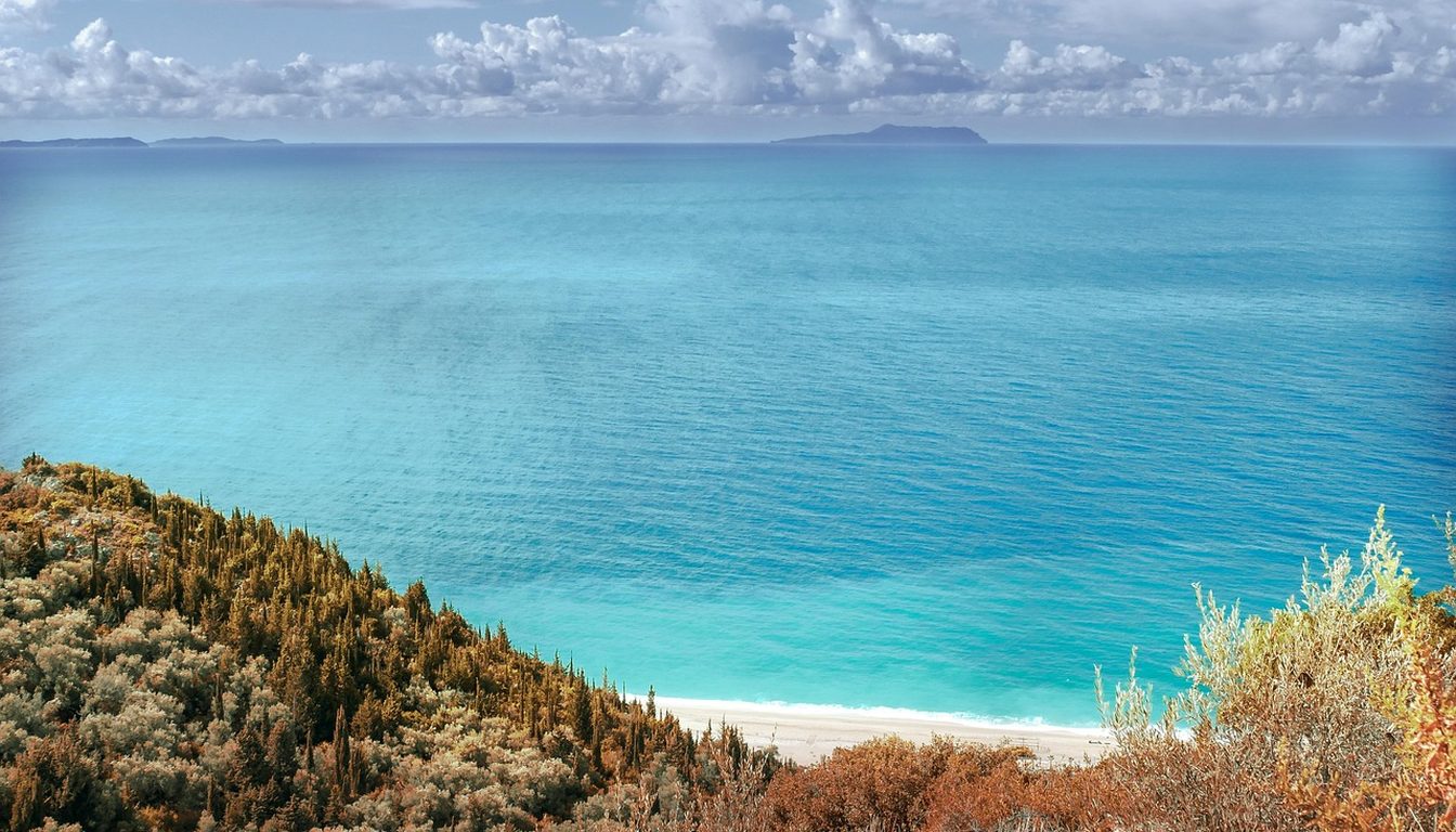 Empty white sand beach below scrub-covered hillside, with turquoise sea stretching to distant islands