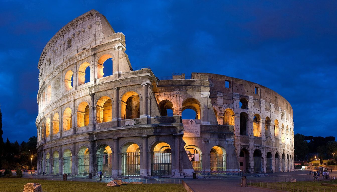 The Colosseum lit up at dusk under a deep blue sky, its arched tiers glowing warmly
