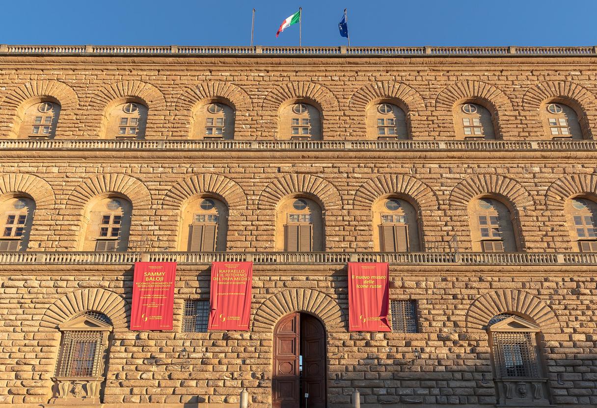 Rusticated stone facade of Palazzo Pitti with arched windows, red exhibition banners and flags under a blue sky