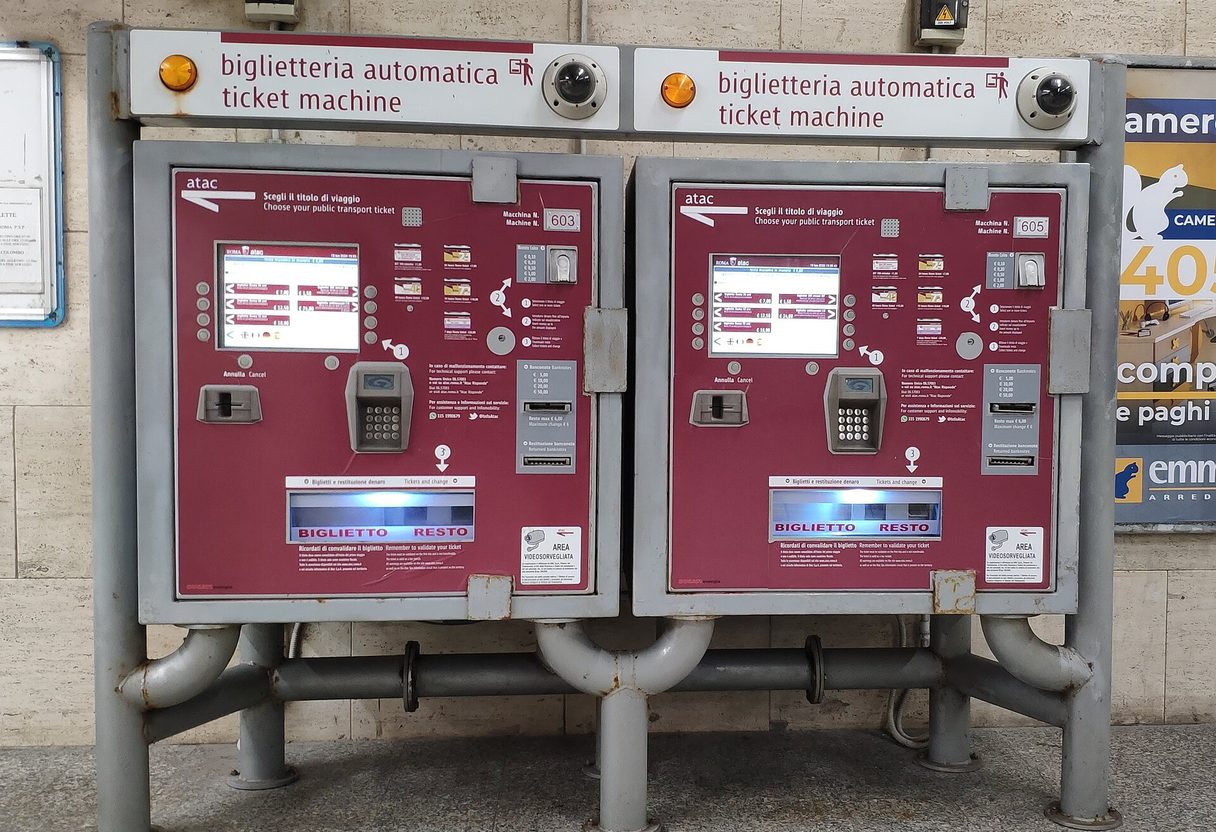 Two red ATAC ticket vending machines with touchscreens and card keypads mounted on a metal stand in Rome