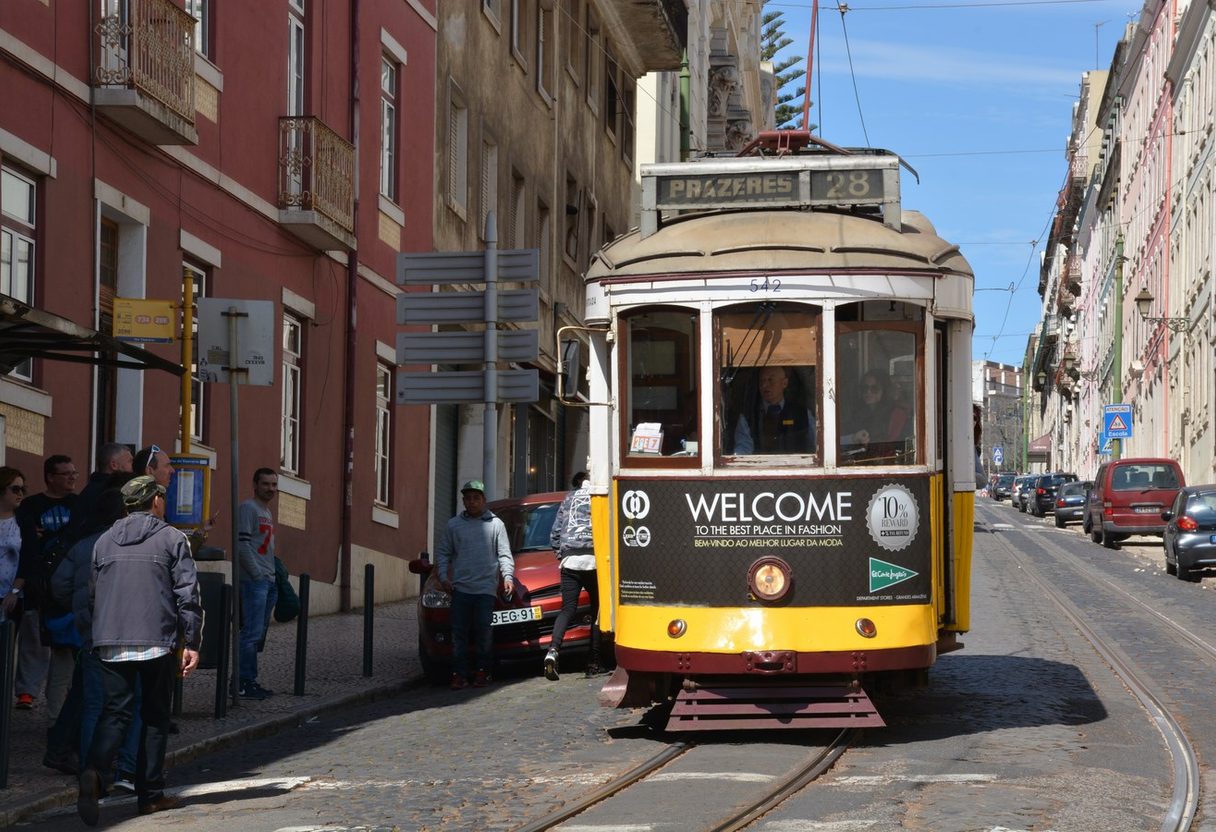 Tram 28 to Prazeres on a cobbled Alfama street with pedestrians and parked cars