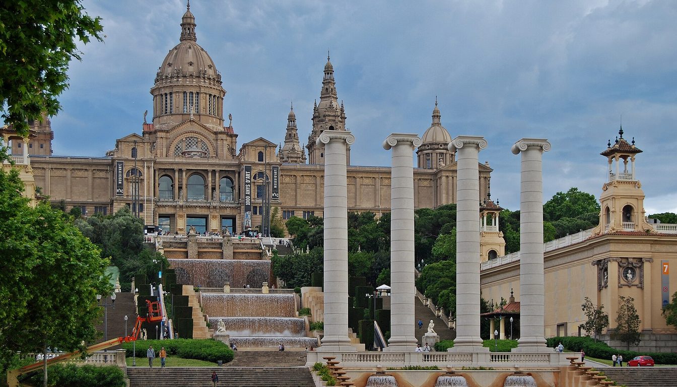 Palau Nacional on Montjuïc above cascading fountains and steps, with the Four Columns in front under a cloudy sky