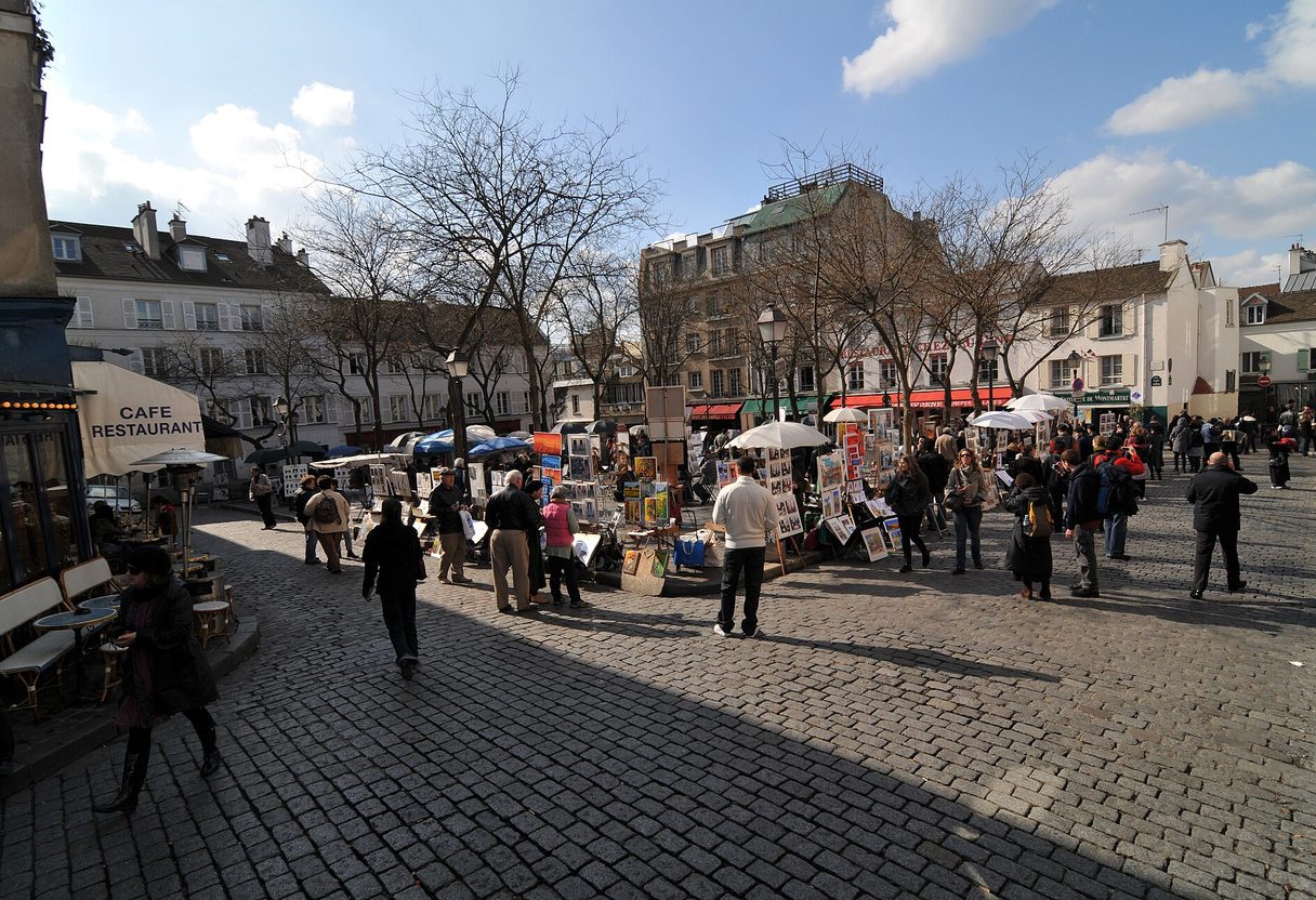 Wide view of cobbled Place du Tertre with painters' stalls, umbrellas, bare trees and café fronts under blue sky