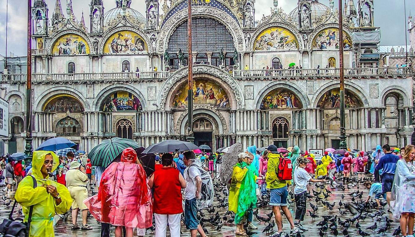 Crowds of tourists in colourful rain ponchos and umbrellas fill a wet St Mark's Square in front of the basilica, with pigeons