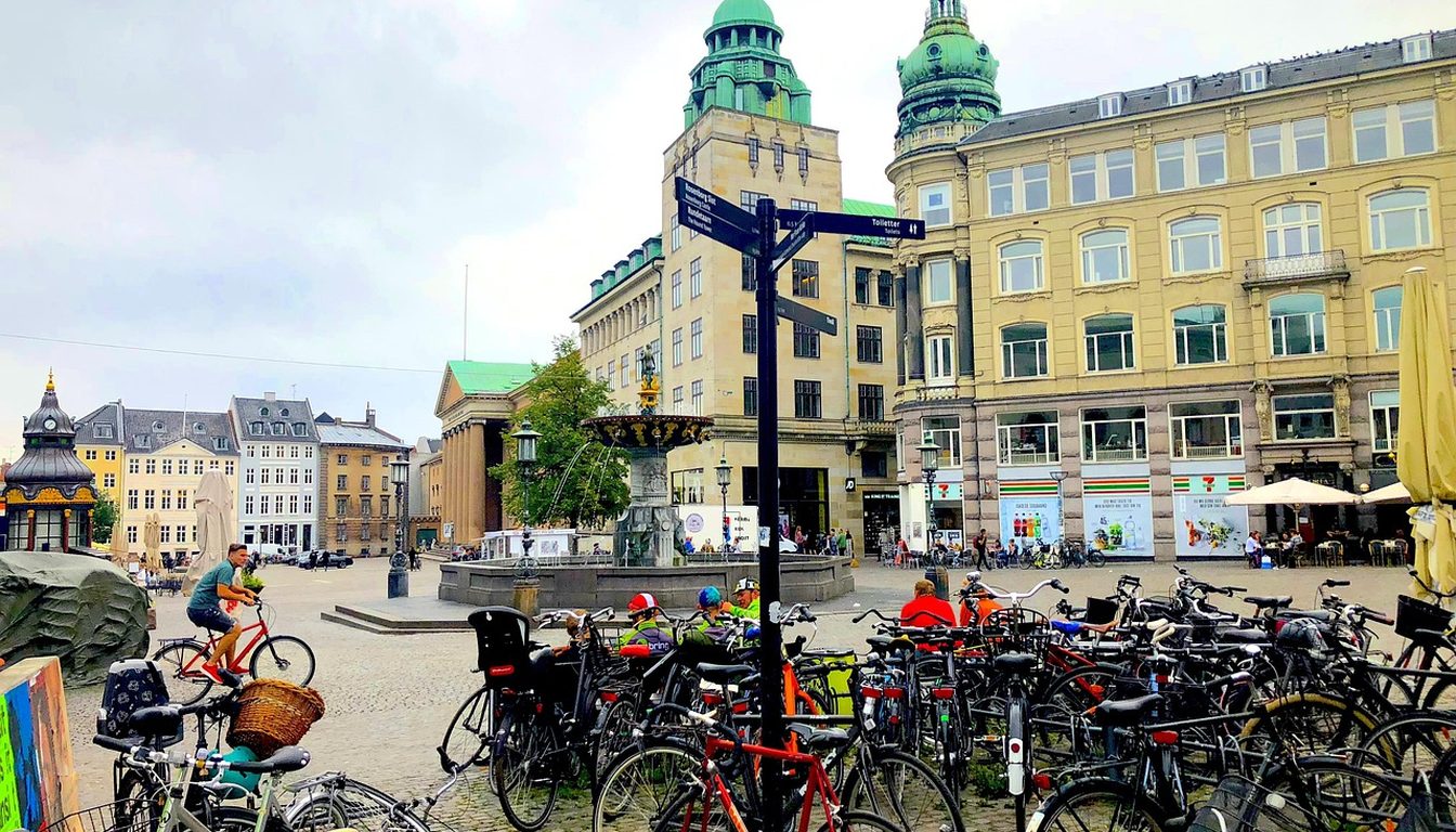 Dozens of bicycles parked on a Copenhagen square with a cyclist passing, a fountain and green-domed buildings behind